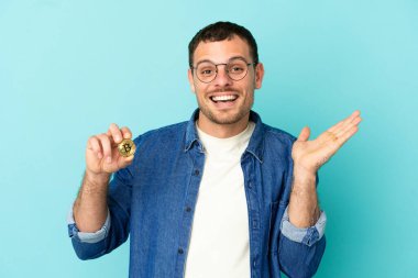 Brezilyalı bir adam Bitcoin 'i izole edilmiş mavi arkaplanda tutuyor ve şok olmuş bir yüz ifadesi var.