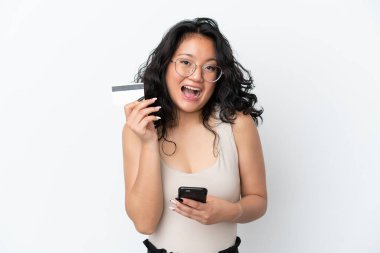 Genç Asyalı bir kadın beyaz arka planda izole edilmiş cep telefonuyla alışveriş yapıyor ve elinde şaşırmış bir ifadeyle bir kredi kartı tutuyor.