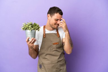 Brezilyalı bahçıvan, mor arka planda bir bitki tutuyor ve gülüyor.