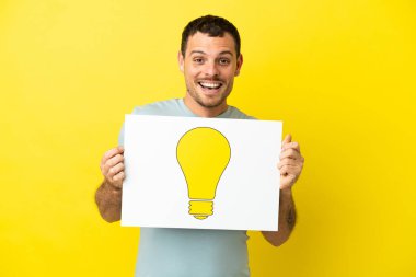 İzole edilmiş mor arka plan üzerinde Brezilyalı bir adam elinde mutlu ifadeli bir ampul simgesi olan bir levha tutuyor.