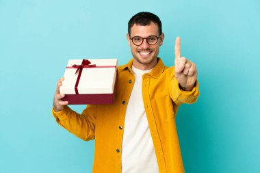 İzole edilmiş mavi arka plan üzerinde bir hediye tutan Brezilyalı adam parmağını kaldırıyor ve gösteriyor.