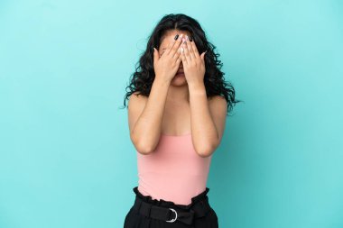 Mavi arka planda izole edilmiş genç Asyalı kadın gözleri elleriyle kapatıyor.