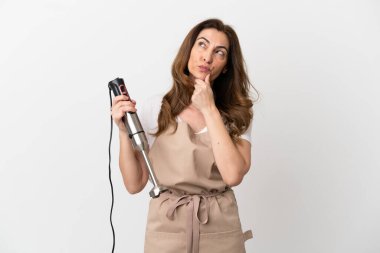 Beyaz arka planda izole edilmiş el blenderi kullanan orta yaşlı beyaz kadın şüpheleri var.