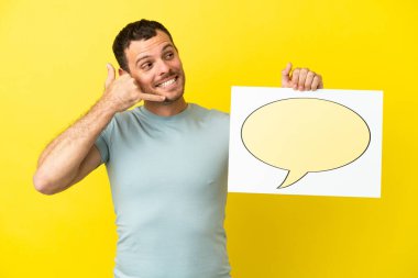 İzole edilmiş mor arka plan üzerinde Brezilyalı bir adam elinde konuşma balonu ikonu olan bir levha tutuyor ve telefon jesti yapıyor.