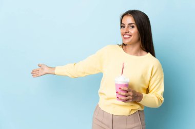 Çilekli milkshake 'li genç Uruguaylı kadın. Mavi arka planda izole edilmiş. Davet etmek için ellerini yana uzatıyor.