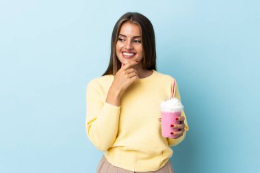 Mavi arka planda izole edilmiş çilekli milkshake 'li genç Uruguaylı kadın yan tarafa bakıyor ve gülümsüyor.
