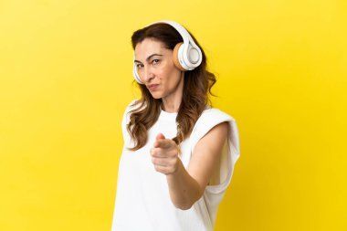 Orta yaşlı beyaz kadın sarı arka planda izole edilmiş müzik dinliyor ve ön tarafı işaret ediyor.