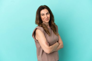 Orta yaşlı beyaz kadın, mavi arka planda izole edilmiş, kolları bağlı ve ileriye bakıyor.