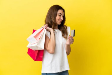 Sarı arka planda izole edilmiş orta yaşlı beyaz kadın elinde alışveriş çantaları ve bir kredi kartı tutuyor.