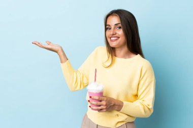 Çilekli milkshake 'li genç Uruguaylı kadın. Mavi arka planda izole edilmiş. Davet etmek için ellerini yana uzatıyor.