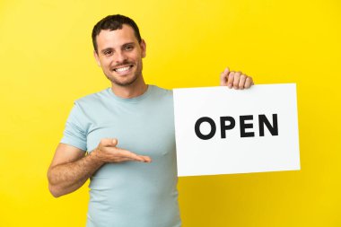 Mor arkaplan üzerinde Brezilyalı bir adam açık yazılı bir pankart tutuyor ve onu işaret ediyor.