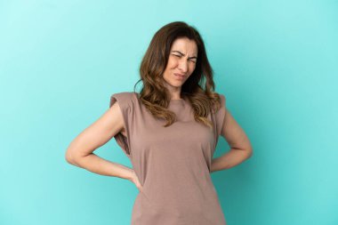 Orta yaşlı, beyaz bir kadın, mavi arka planda izole edilmiş, gayret gösterdiği için sırt ağrısı çekiyor.