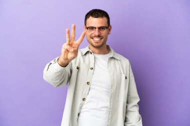 İzole edilmiş mor arka planı olan Brezilyalı adam mutlu ve parmaklarıyla üçe kadar sayıyor.
