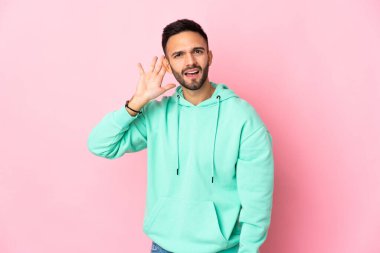 Pembe arka planda izole edilmiş genç beyaz adam elini kulağına koyarak bir şeyler dinliyor.