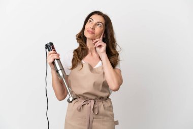Orta yaşlı beyaz kadın el blenderi kullanıyor. Beyaz arka planda izole edilmiş, yukarı bakarken bir fikir düşünüyor.