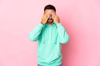 Pembe arka planda izole edilmiş genç, beyaz bir adam gözlerini elleriyle kapatıyor.