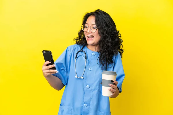 Young surgeon doctor asian woman isolated on yellow background holding coffee to take away and a mobile