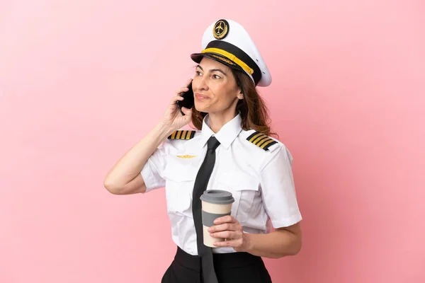 Airplane middle aged pilot woman isolated on pink background holding coffee to take away and a mobile