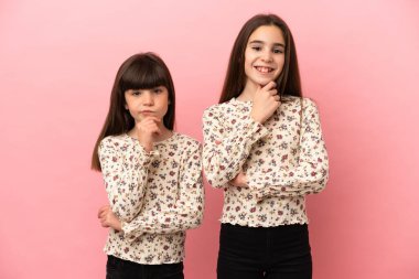 Küçük kız kardeşler pembe arka planda izole edilmiş gülümseyen ve kendinden emin bir yüzle öne bakan kızlar.