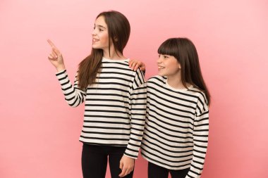 Pembe arka planda izole edilmiş küçük kız kardeşler bir ürün sunmak için kenarı gösteriyorlar.