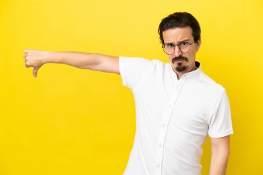 Sarı arka planda izole edilmiş genç beyaz adam olumsuz ifadelerle baş parmağını aşağı gösteriyor.