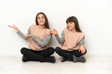 Küçük kız kardeşler beyaz arka planda izole bir şekilde yerde oturuyor. Davet edilmek için ellerini yana uzatıyor.