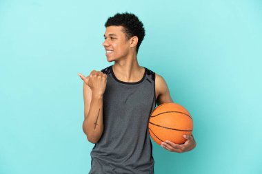 Basketbolcu Afrikalı Amerikalı bir adam mavi arka planda izole edilmiş bir şekilde bir ürünü sunmak için kenarı işaret ediyor.