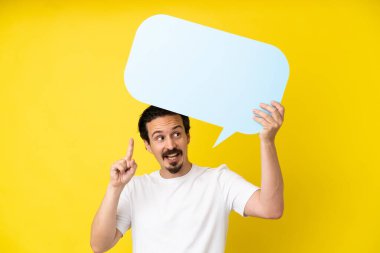 Sarı arka planda izole edilmiş genç beyaz adam boş bir konuşma balonu tutuyor ve düşünüyor.