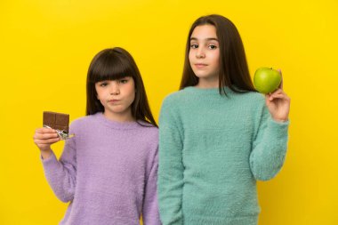 Sarı arka planda izole edilmiş küçük kız kardeşler bir elinde çikolata tableti, diğer elinde elma tutarken şüpheye düşüyorlar.
