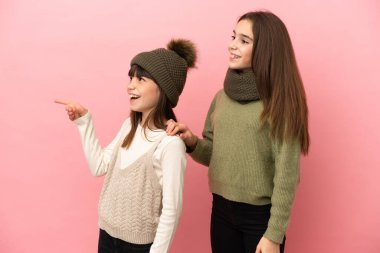 Pembe arka planda izole edilmiş kışlık giysiler giyen küçük kardeşler bir ürünü sunmak için kenarı gösteriyorlar.