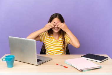 Mor arka planda dizüstü bilgisayarı olan küçük bir öğrenci kız. Gözlerini elleriyle kapatıyor ve gülümsüyor.