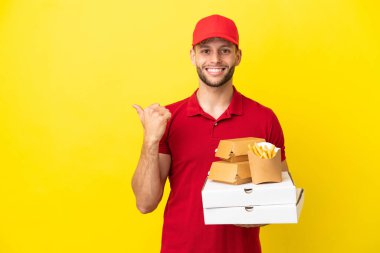 Pizza teslimatçısı, izole edilmiş arka planda pizza kutularını ve hamburgerleri topluyor. Ürünü sunmak için yan tarafa bakıyor.