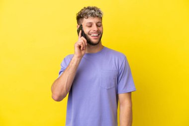 Sarı arka planda izole edilmiş cep telefonu kullanan genç beyaz adam gülüyor.