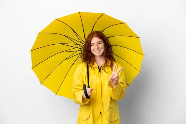 ティーンRedhead女の子雨防止コートと傘隔離上の白い背景笑顔とショー勝利サイン — ストック写真