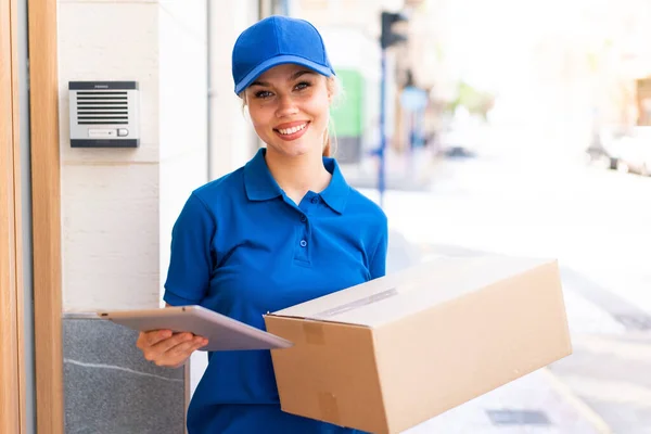 Young delivery woman at outdoors holding boxes and a tablet with happy expression