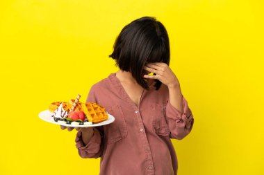 Pregnant woman holding waffles isolated on yellow background with tired and sick expression