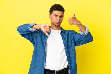 Genç Afro-Amerikalı adam sarı arka planda izole edilmiş, iyiye-kötüye işaret yapıyor. Evet ya da hayır arasında kararsız