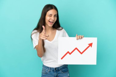 Genç Fransız kadın mavi arka planda izole edilmiş. Elinde büyüyen bir istatistik oku sembolü olan bir tabela var ve ön tarafı işaret ediyor.