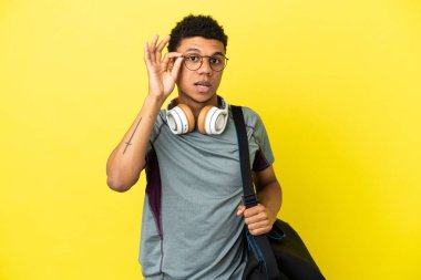 Spor çantalı genç Afrikalı Amerikalı adam sarı arka planda gözlüklü ve şaşırmış.
