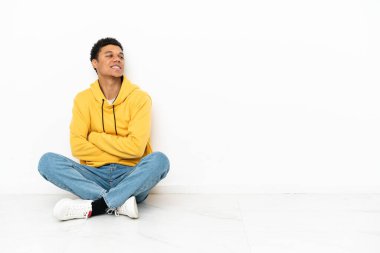 Young African American man sitting on the floor isolated on white background with arms crossed and happy
