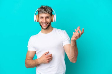 Genç yakışıklı beyaz adam mavi arka planda izole edilmiş müzik dinliyor ve gitar hareketi yapıyor.