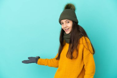 Mavi arka planda izole edilmiş genç bir Fransız kız. Kış şapkası takıyor. Davet etmek için ellerini yana açıyor.