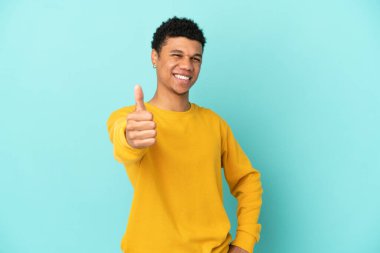 Genç Afrikalı Amerikalı adam mavi arka planda izole edildi. Başparmakları havada çünkü iyi bir şey oldu.