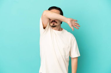 Genç beyaz adam mavi arka planda izole edildi. Gözlerini elleriyle kapadı.