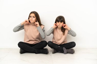 Küçük kız kardeşler beyaz arka planda izole bir şekilde yerde oturuyorlar. İki kulağı da elleriyle kaplıyorlar.