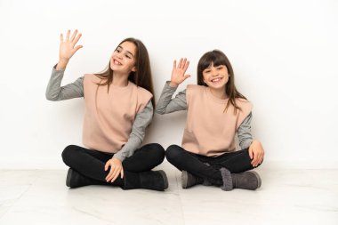Beyaz arka planda izole edilmiş yerde oturan küçük kız kardeşler mutlu bir ifadeyle selamlaşıyorlar.