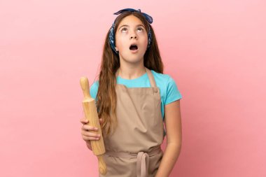 Küçük beyaz kız pembe arka planda izole edilmiş bir oklavayı tutuyor yukarı bakıyor ve şaşırmış bir ifadeyle