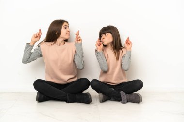Küçük kız kardeşler beyaz arka planda izole bir şekilde yerde oturuyor parmaklarını çapraz yapıp en iyisini diliyorlar.