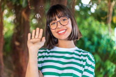 Parkta genç esmer bir kadın, mutlu bir ifadeyle selamlıyor.