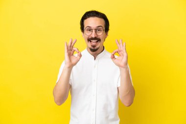 Sarı arka planda izole edilmiş genç beyaz adam iki eliyle tamam işareti gösteriyor.